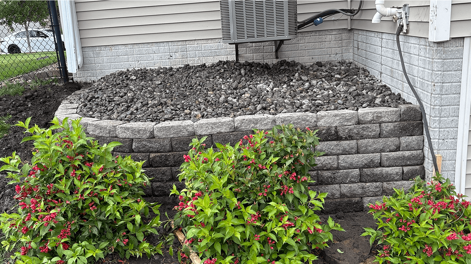 Retaining wall installation with black rock and flowering plants around home foundation.